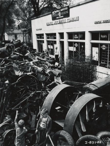 Two young boys in a pile of metal collected from a scrap drive in front of the Farmers Mercantile Company, an International Harvester dealership. A man stands near the entrance of the building.