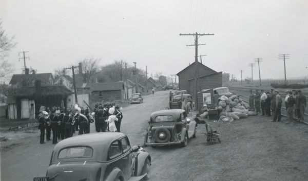 The Elk Mound High School band entertaining farmers as they weigh in loads of scrap at Ausman Implement Company, an International Harvester dealership. The metal was collected for a scrap drive. In the foreground cars are lined up, and a group of men stand near railroad tracks.