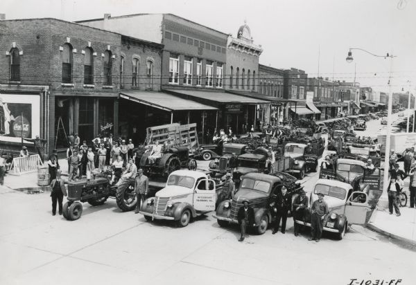 Elevated view of a large group of people gathered with tractors and trucks for a scrap parade in a downtown area. In the background automobiles are parked at an angle in front of storefronts.
