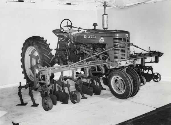 Farmall Super M-TA tractor with mounted plow and cultivator unit on display in front of a white backdrop. The equipment may be experimental.