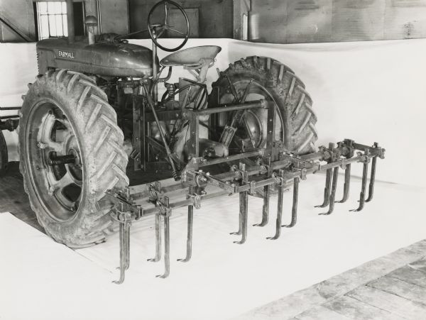 Farmall tractor with an H&M tool bar and diamond point cultivators on display in front of a white backdrop. The equipment may be experimental.