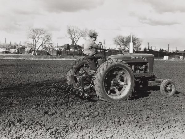 Right side view of a man driving a Farmall M tractor with mounted H&M tool bar with 9 coil shanks in a field.