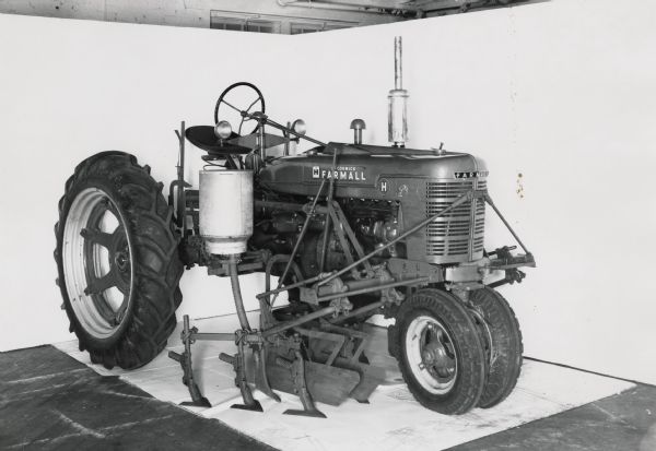 Farmall H tractor with a mounted HM-238 cultivator with a large capacity Richmond style fertilizer attachment. The machinery is on display in front of a white backdrop and may be experimental.