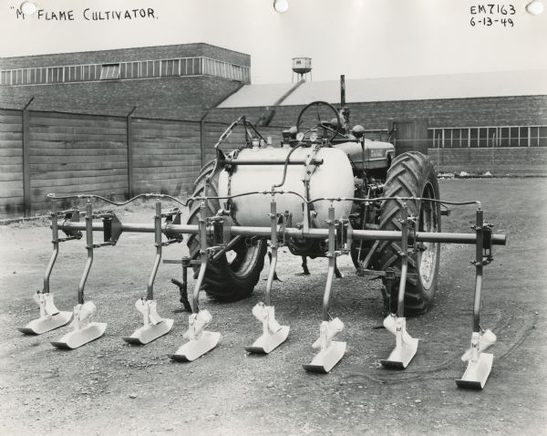 Engineering photograph of a Farmall M(?) tractor with an "M" flame cultivator in a yard outside an International Harvester factory. The machinery may be experimental.