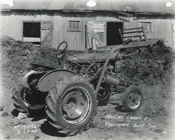 Three-quarter view from right rear of a Farmall A tractor with mounted manure loader and pneumatic lift-all outside farm building or factory test site. The machinery may be experimental.