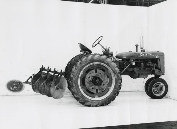 Right side profile view of a harrow plow for 2 point hitch attached to a Farmall Super C tractor. The machinery was photographed against a white backdrop and may be experimental.
