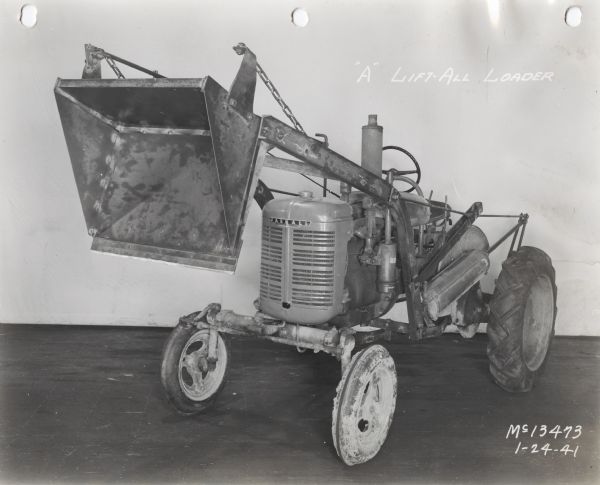 Three-quarter view from front left of "A" lift-all loader mounted on a Farmall A tractor. In the background is a white wall for a backdrop. The equipment may be experimental.
