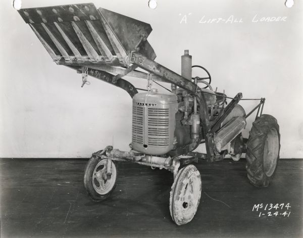 Three-quarter view from left front of "A" lift-all loader mounted on a Farmall tractor. In the background is a white wall for a backdrop. The equipment may be experimental.