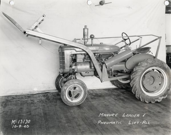 Pneumatic lift-all and manure loader mounted on a Farmall A tractor on display in front of a white curtain backdrop. The equipment may have been experimental.