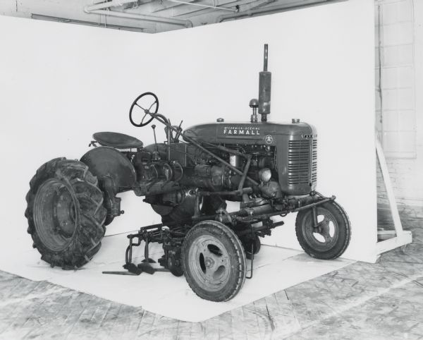 Three-quarter view from right front of an A-647 vegetable cultivator attached to a Farmall A tractor. In the background is a white wall for a backdrop. The equipment may be experimental.