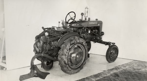 Three-quarter view from right rear of a Farmall A tractor equipped with a moldboard plow. In the background is a white wall for a backdrop. The equipment may be experimental.