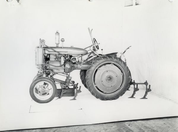 Left side profile view of a Farmall A tractor equipped with an AV-138 cultivator with pneumatic lift. In the background is a white wall for a backdrop. The equipment may be experimental.