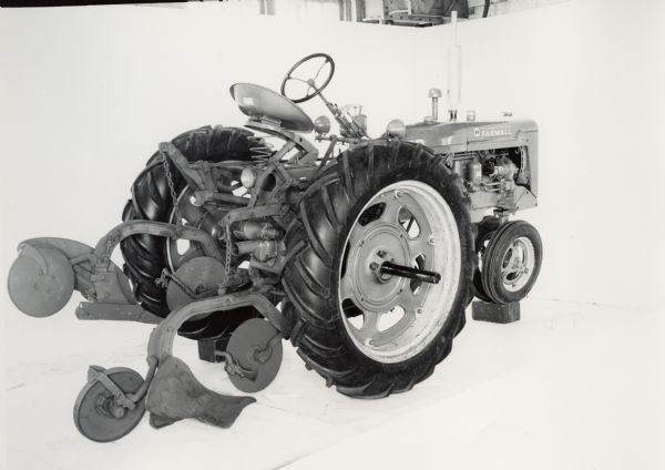 Three-quarter view from right rear of a Farmall C tractor equipped with a No. 199 plow with jointer and furrow wheel at rear. In the background is a white wall for a backdrop. The equipment may be experimental.