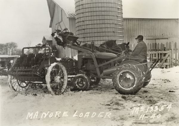 Man using a Farmall A tractor with loader to load manure in 10 degree weather outside a silo and farm building. Two horses are pulling the manure spreader.