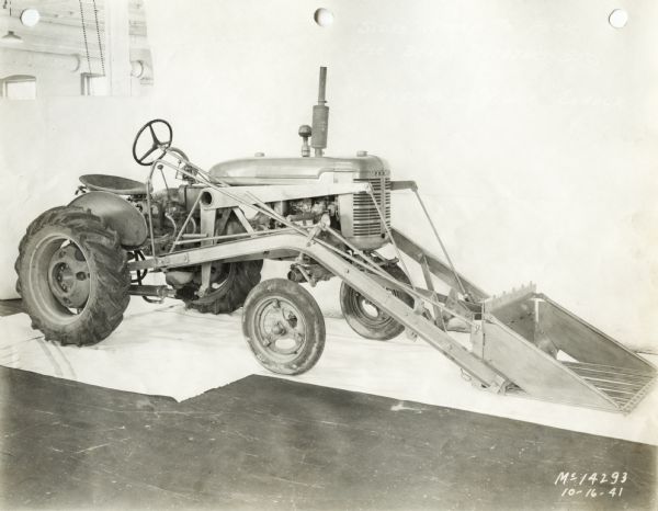 Three-quarter view from right front of a Farmall A tractor with mounted loader attachment. In the background is a white wall for a backdrop. The equipment may be experimental.
