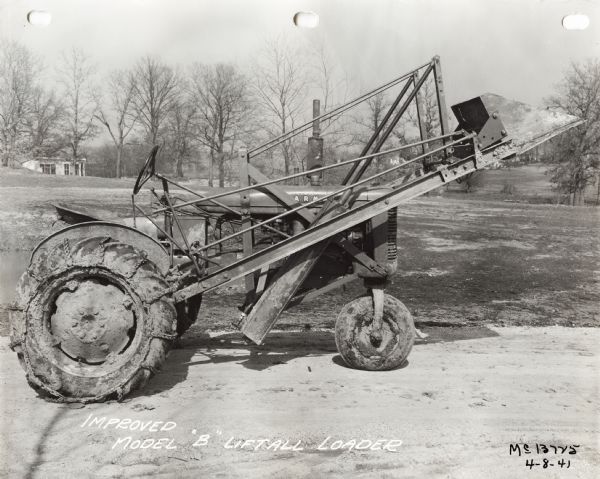 View of right side of a Farmall B(?) tractor equipped with rear tire chains and an "improved model B" liftall loader. The tractor is parked outdoors near a pond.
