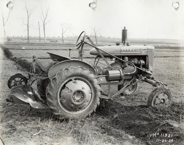 Right side profile view of a Farmall A tractor on farmland with a rear-mounted plow.