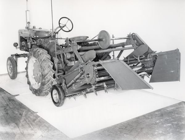 Three-quarter view from left rear of a Peanut shaker attachment mounted at rear of Farmall A tractor. In the background is a white wall for a backdrop. The equipment may be experimental.
