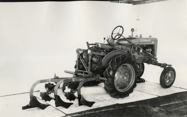 Three-quarter view from right rear of a Farmall A Vineyard plow attached to a Farmall A tractor. In the background is a white wall for a backdrop. The equipment may be experimental.