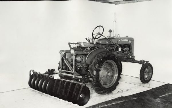 Three-quarter view from right rear of a Farmall A tractor with disk harrow attachment. In the background is a white wall for a backdrop. The equipment may be experimental.