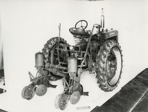 Three-quarter view from right rear of a Farmall C tractor equipped with a No. 46 lister. In the background is a white wall for a backdrop. The equipment may be experimental.