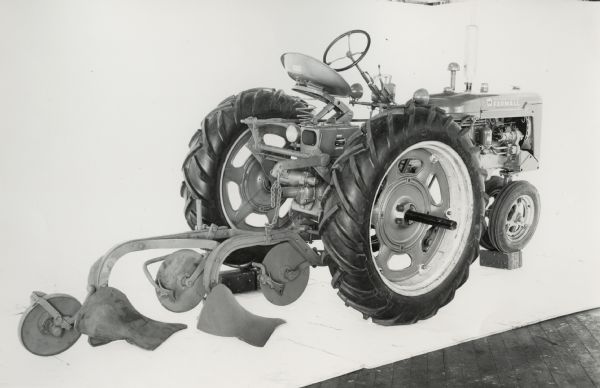 Three-quarter view from right rear of a Farmall C tractor equipped with a No. 295 plow with jointer and furrow wheel. In the background is a white wall for a backdrop. The equipment may be experimental.
