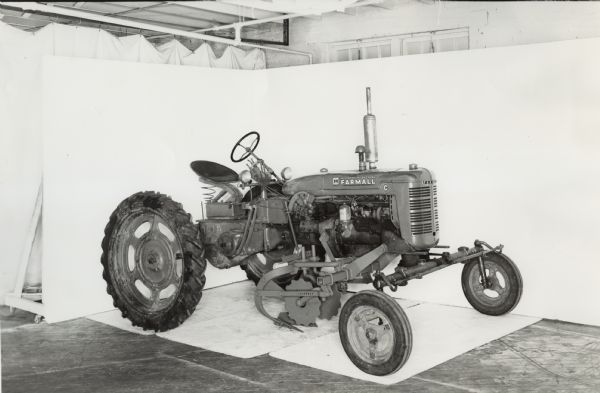 Three-quarter view from front of right side of a Farmall C tractor equipped with No. 8 beet puller. In the background is a white wall for a backdrop. The equipment may be experimental.