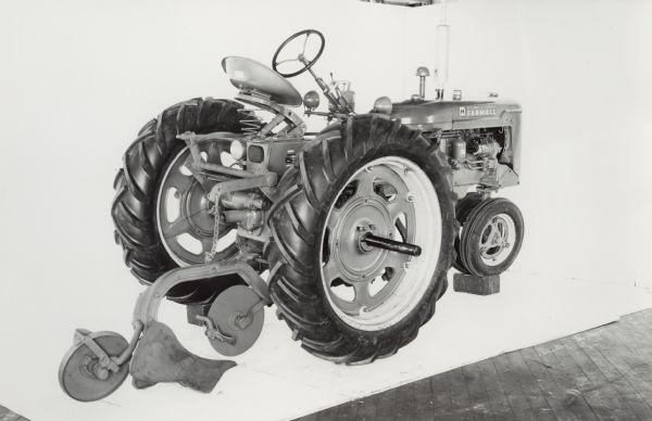 Engineering three-quarter view from right rear of a Farmall C tractor equipped with plow with jointer and furrow wheel at rear. In the background is a white wall for a backdrop. The equipment may be experimental.