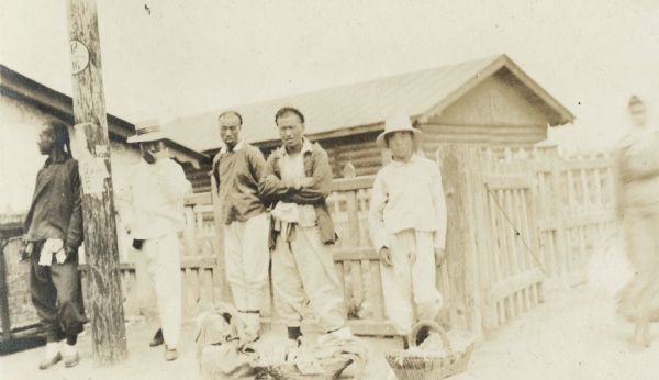 Group of "Chinese laborers" standing along wooden fence outside small building. In front of the men are baskets on the ground.