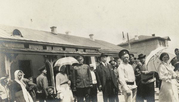 Group of men, women and children outside the train station at Hailar in Manchuria. Two women hold umbrellas for protection from the sun. The original caption identifies two of the men as "Col. Michie" and "Mr. Duncan."