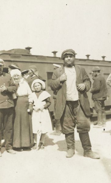A bearded man wearing a hat and boots is standing near a passenger train. On the left is a woman with a young girl looking on. The original caption reads: "a Russian peasant."