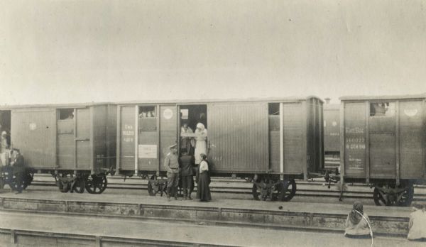 Men and women wait on and near box cars at a train platform in Russia. The original caption reads: "cheapest traveling on the Trans-Siberian Ry. [Railway]."