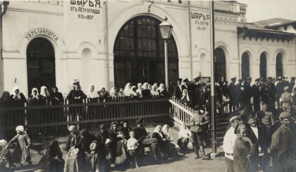 Crowd of women, children and soldiers at a train station on the Trans-Siberian Railway. The station may have been located between Mogzon and Vyatka, Russia. The original caption reads: "giving the Mission the 'once over.'" A caption for a similar view reads: "peasant women and children at station in European Russia."