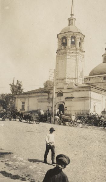 Horse-drawn wagons outside a large church with belfry in a town along the Trans-Siberian Railway. The original caption reads: "tower of 300 year old church at Vyatka."
