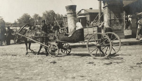 Horse pulling a woman on a wagon in a town along the Trans-Siberian Railway. The woman is sitting in a large wicker basket, and behind her is a box tied with a rope. A man in a uniform and hat is standing behind the wagon, and in the background are posters on a column. The original caption reads: "peasant woman at Vyatka."