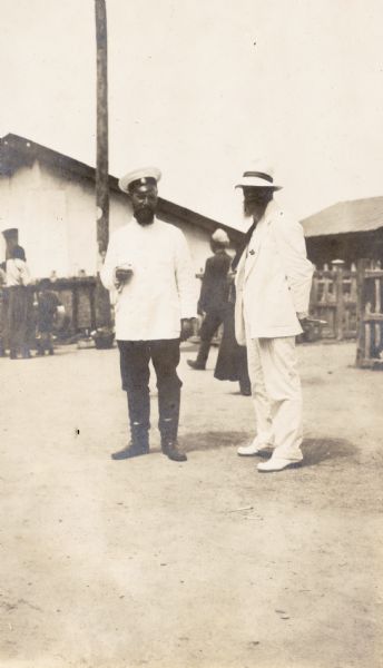 Man dressed in white suit standing outside with uniformed man. Original caption reads: "Russian station master and wealthy peasant at Manchuria."