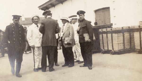 Men standing near a building, probably at a train station, in Manchuria. There are a number of large baskets on wheels sitting outside of a door of the building. The original caption reads: "Mr. Gorbatenko and Russian officials at Manchuria."