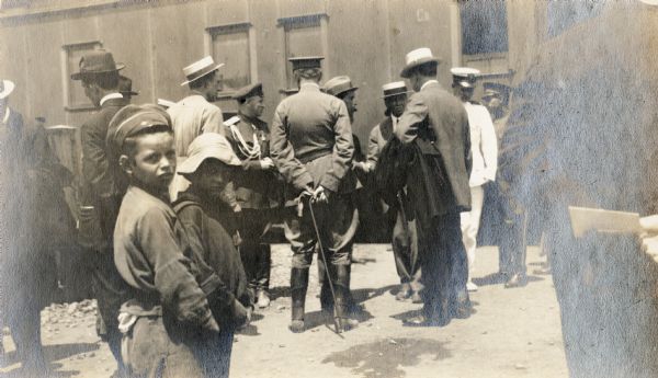 Several well-dressed men stand near uniformed men outside a railroad car. Two young boys are in the foreground looking back at the photographer. Original caption reads: "arrival at Vladivostok."