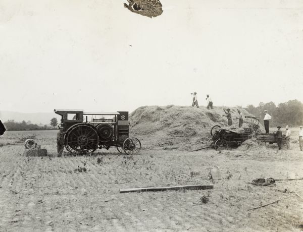 Several men baling hay into hay press belted to a Mogul 15-30 or 30-60 tractor for power. Several other men supervise nearby. A barrel of oil sits behind the men on a bale of hay.