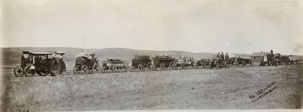 Panoramic view of a Mogul 30-60(?) tractor pulling a caravan of wagons loaded with various goods. Men are standing on and around the tractor and wagons.