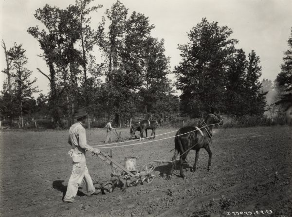 A man using a P&O walking planter pulled by one horse in a field. In the background another man works using a walking plow pulled by two horses near a row of trees.