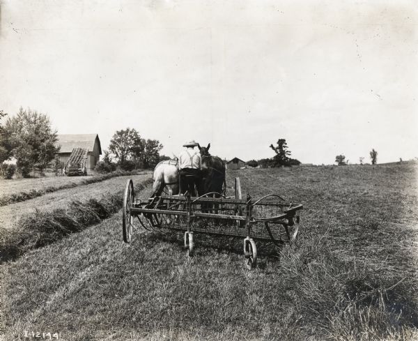 Rear view of man wearing a hat and suspenders using two horses to operate an Osborne side-delivery hay rake, or dump rake in a farm field.  Several barns and farm buildings, and what appears to be a windrow hay loader, are in the background.