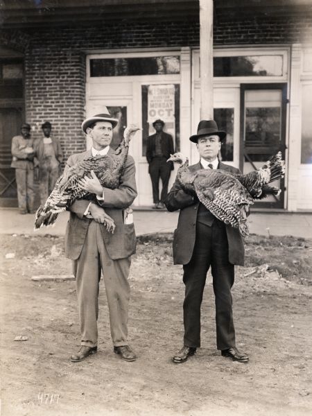 J.M. Browning and Mr. Powell, wearing hats, suits, and ties, each holding a turkey while standing on a dirt road. Several individuals are in the background standing against what appears to be a storefront.