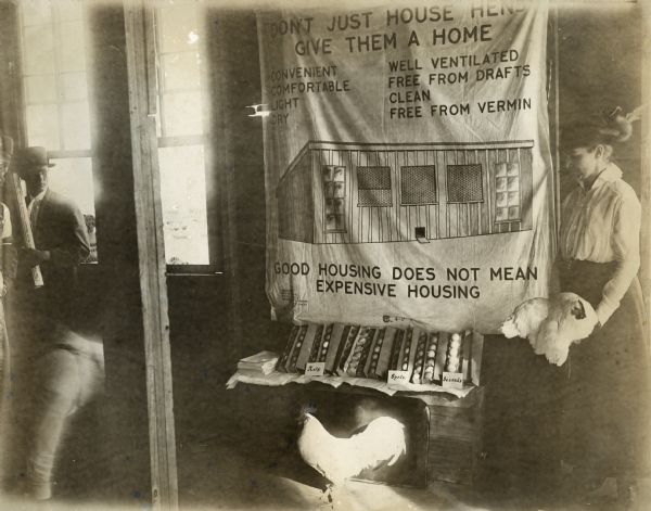 A woman holding a chicken is standing beside a display illustrating quality poultry housing (chicken coops).