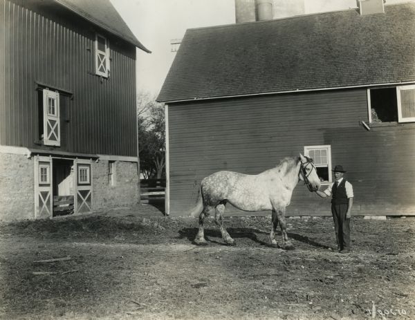 A man wearing a hat and vest stands in front of two farm buildings with his hand on the bridle of what appears to be a dapple gray horse.