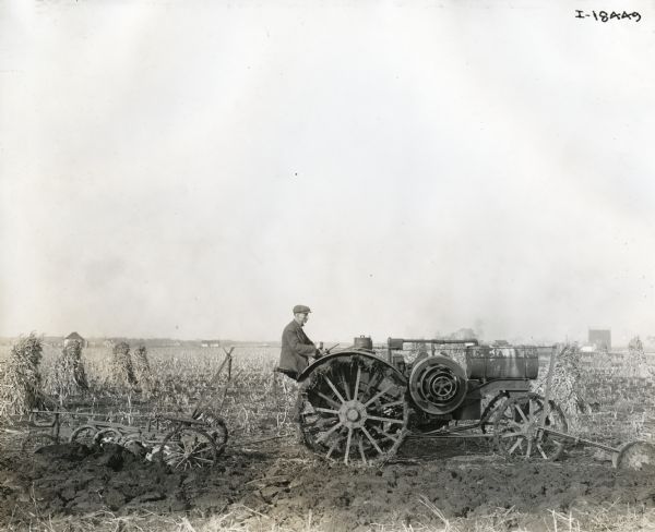 Side view of man using an International Harvester Titan 10-20 tractor and a Janesville three bottom plow to work in a field. Farm buildings are in the far distance.