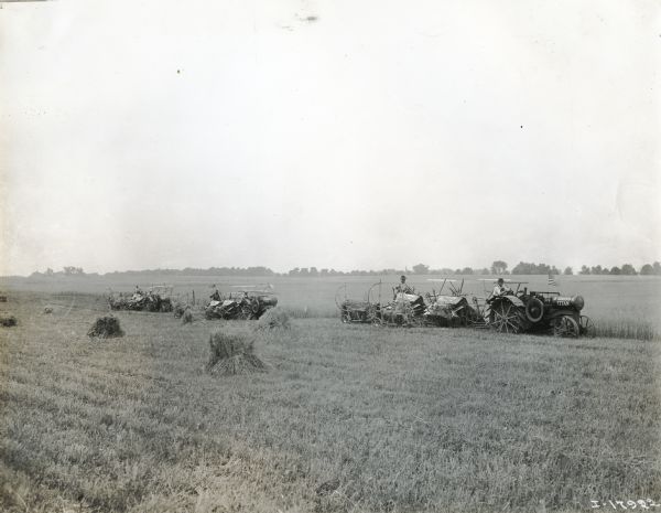 View across field towards six men harvesting what appears to be wheat using International Titan 10-20 tractors and combines (harvester-threshers).