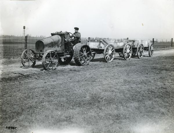 A man is pulling an eight-wheeled wagon down a dirt road using an International 8-16 tractor. A fence and water tower are in the background.