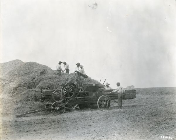Men are standing on a large pile of hay using pitchforks to load hay into a hay press operated by two other men.