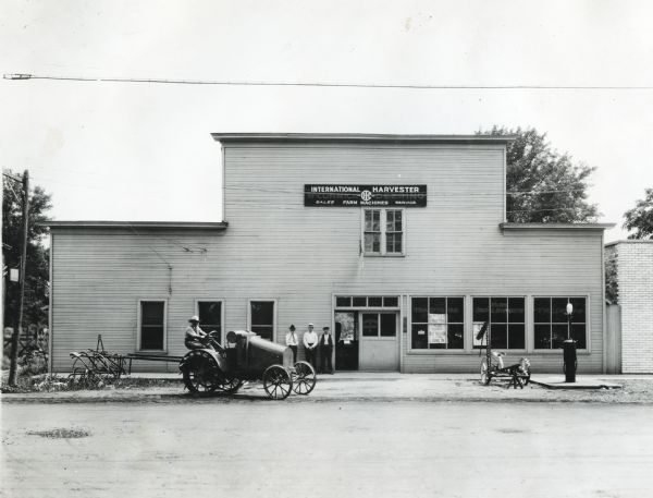 Exterior view of Hayden & Harms, an International Harvester dealership. Several men are standing near the building's front door near displayed farm implements, while another man is sitting behind the wheel of an International 8-16 tractor.